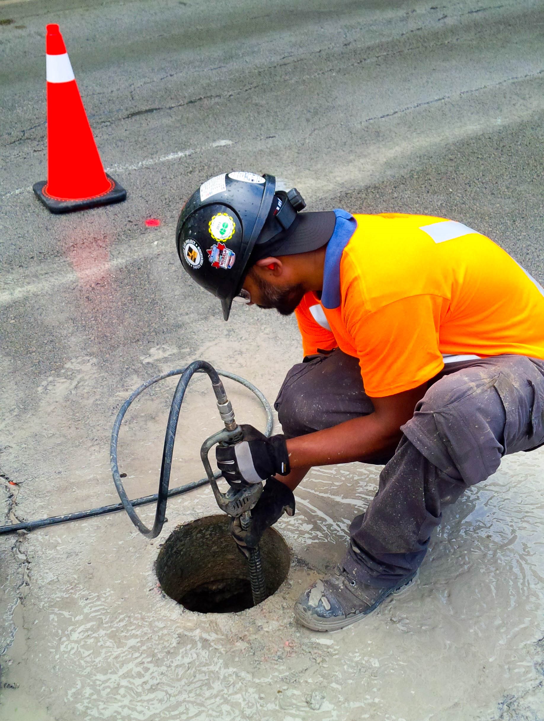 Informative Image displays a multiVIEW field technician kneeled next to a hole in the paved road. The pavement has been recently cored to create an opening (hole). He is bent over using a high pressure water jet to loosen the material covering the target, which is typical of a Hydro Excavation.