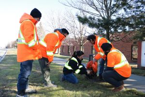 Informative Image showcasing a field supervisor providing training to a team of new utility locators. The field supervisor is kneeling next to a communication box with four new recruits standing around her listening and observing her instructions.